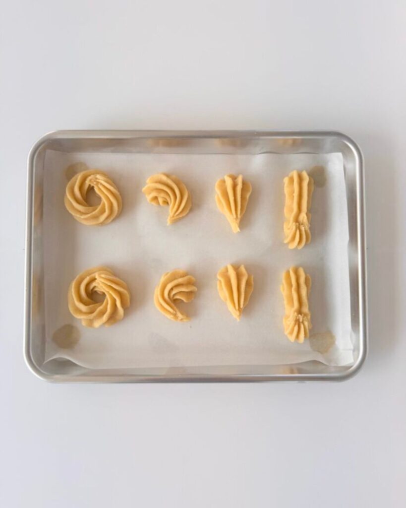 Almond shortbread cookies piped into wheels, shells, and sticks on a parchment-lined sheet pan, ready to bake.