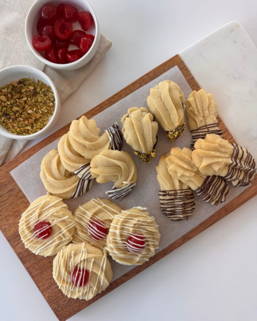 Finished almond shortbread cookies on a wooden board, decorated with white chocolate, dark chocolate, chopped pistachios, and maraschino cherries.