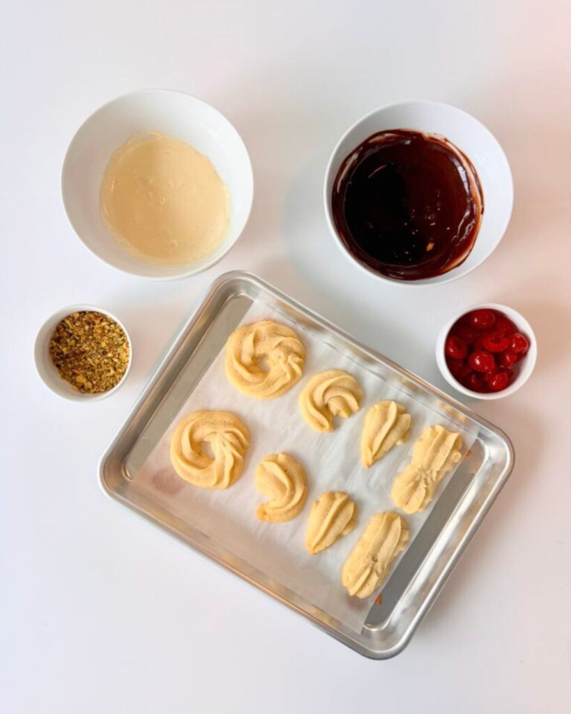 A sheet pan of baked almond shortbread cookies beside bowls of melted white chocolate, dark chocolate, chopped pistachios, and maraschino cherries for decorating.